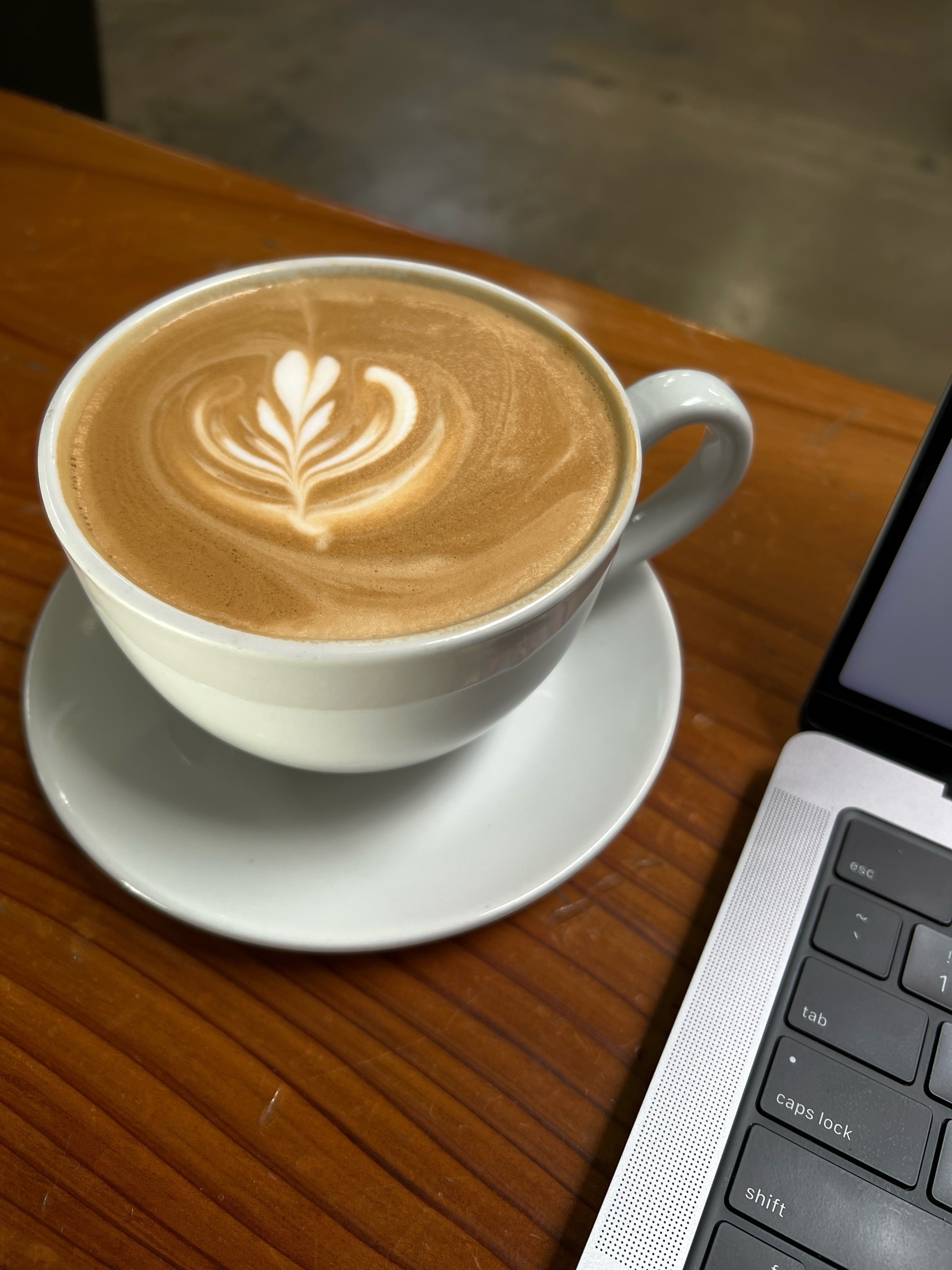a latte in a white ceramic cup, complete with latte art leaf design. a corner of a laptop keyboard appears on the right edge of frame. 