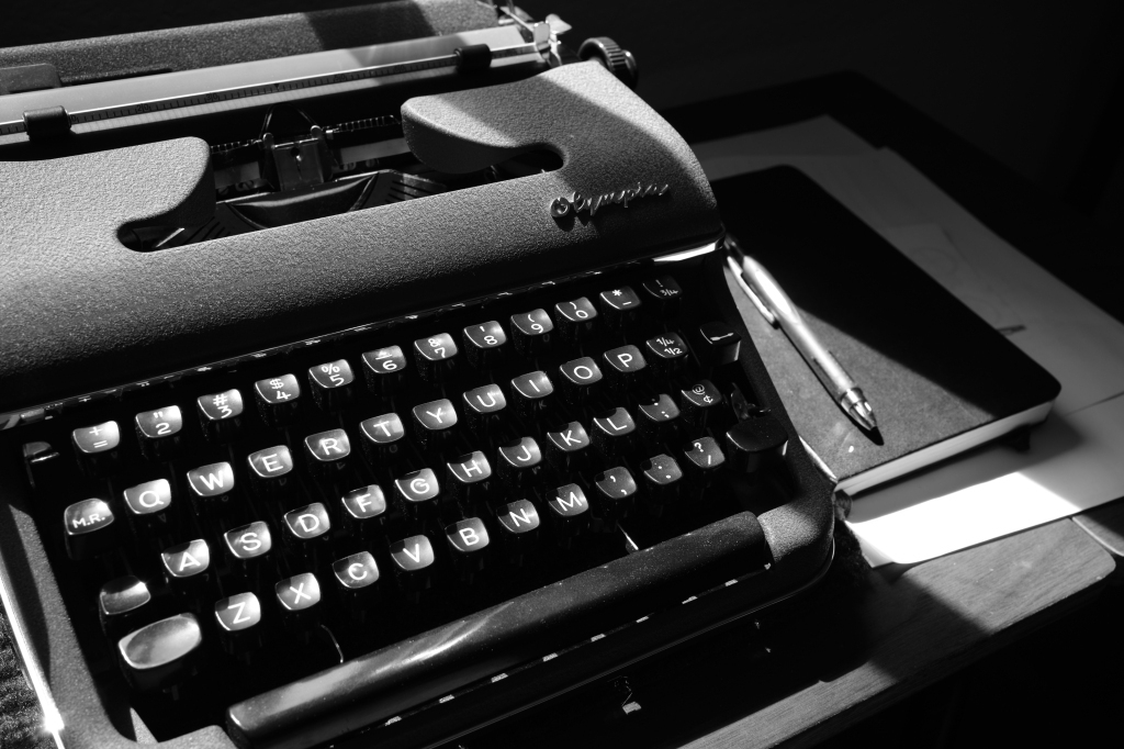 a black and white photo of a Olympia SM2 typewriter, some paper, a notebook, and a pen on a typewriter desk. There is some sunlight on the typewriter, coming from a window that isn't in frame. 