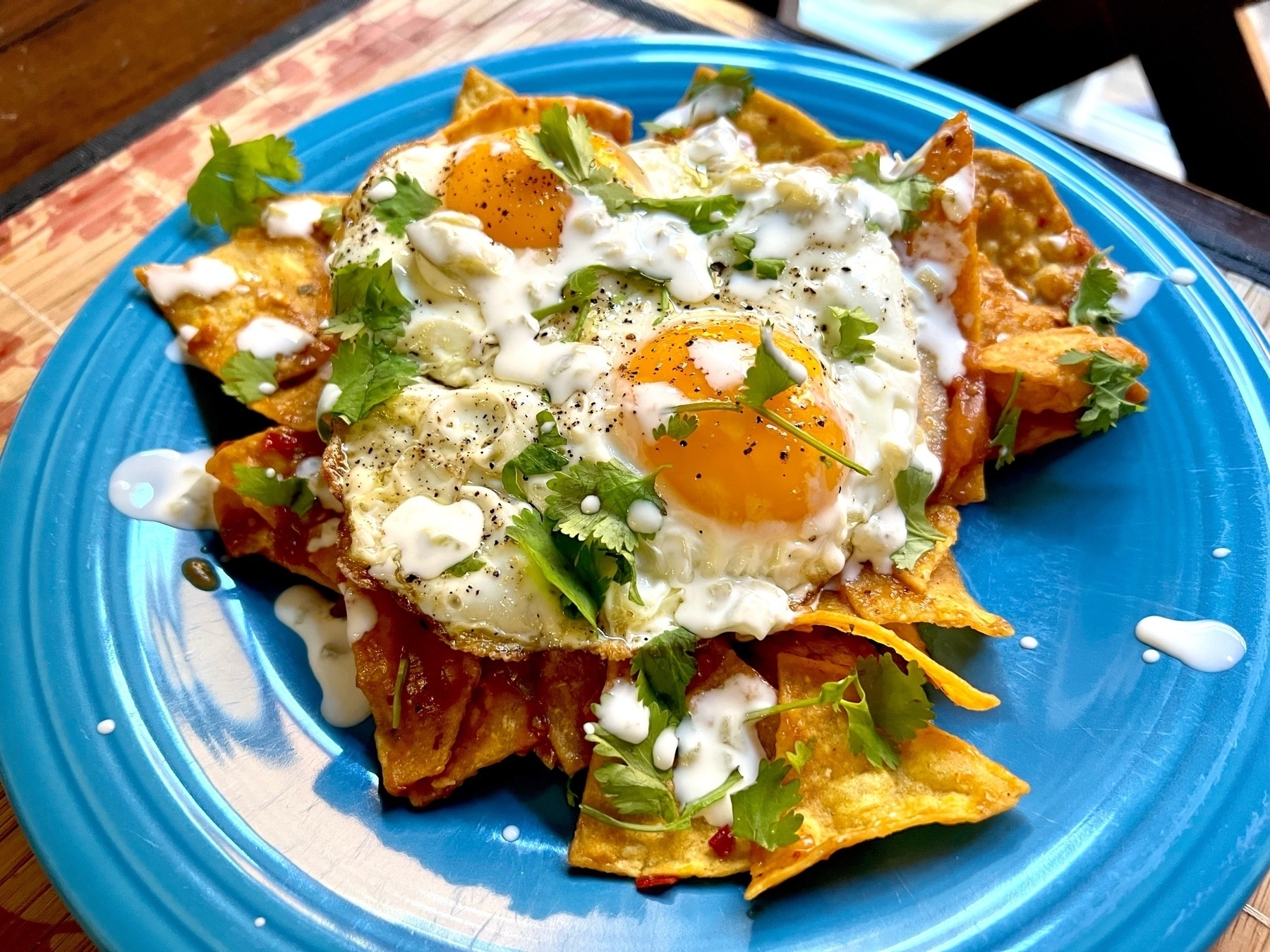 a serving of chilaquiles on a blue plate. corn tortilla chips lightly simmered in salsa, with two fried eggs served sunny side up on top. Sprinkled with chopped cilantro.  
