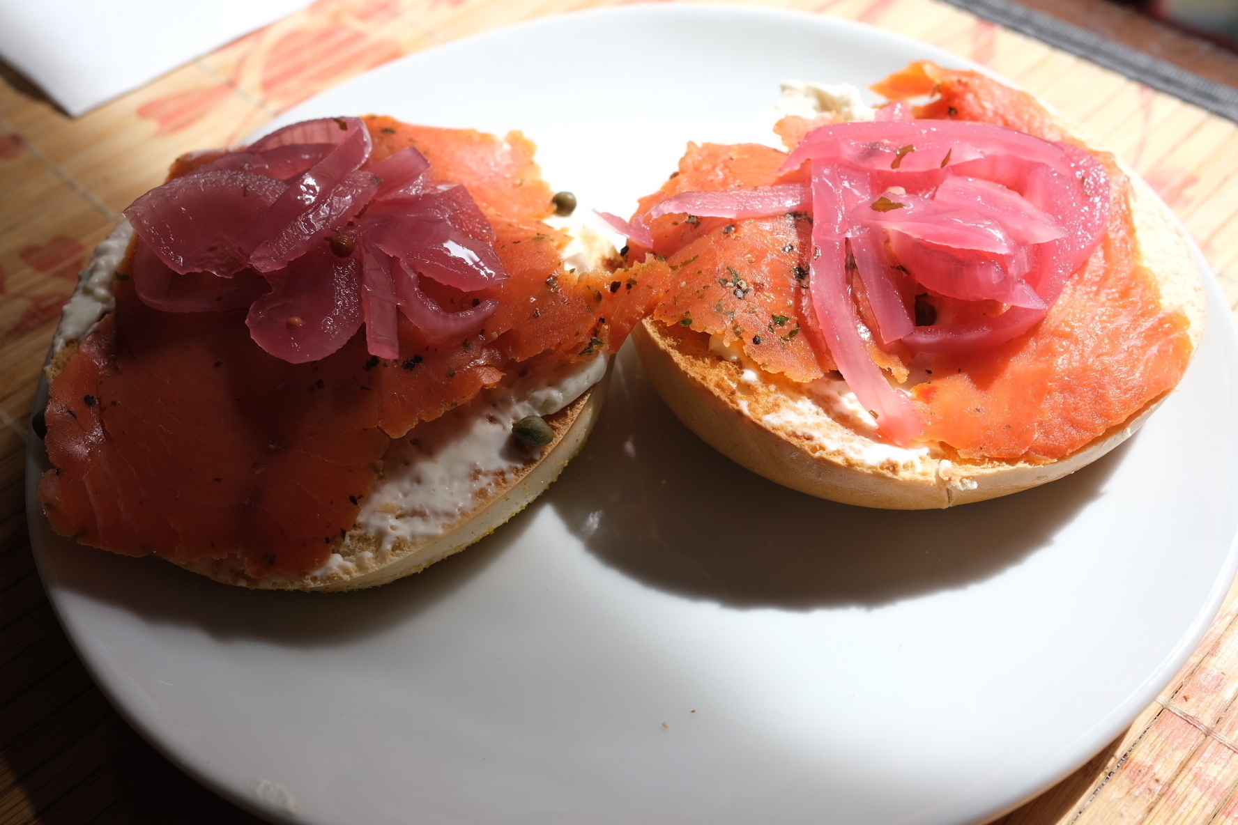 A bagel on a plate with cream cheese, smoked salmon, and pickled onions. Half of it is lit by the morning sunlight coming through a nearby window. 